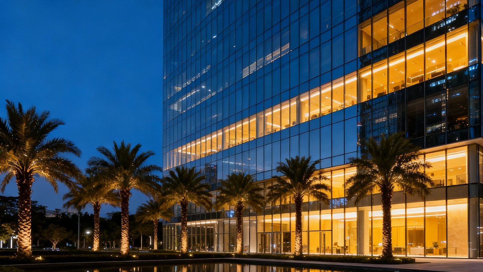 一张展示现代化超甲级写字楼玻璃幕墙外立面的夜景摄影照片，建筑内部暖色灯光与深蓝天幕形成对比，楼前有精致水景和棕榈树，彰显高端商务氛围